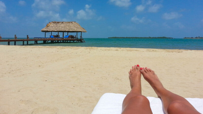 A long strech of sand on one of the best beaches in Belize with a woman's legs on a beach char