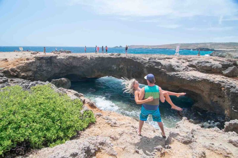 Man holding a woman in front of the Natural Bridge in Aruba - couple travel tips
