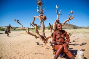 A Himba Tribe Woman sitting in front of a dead tree in the Namib Desert - things to do in Namibia