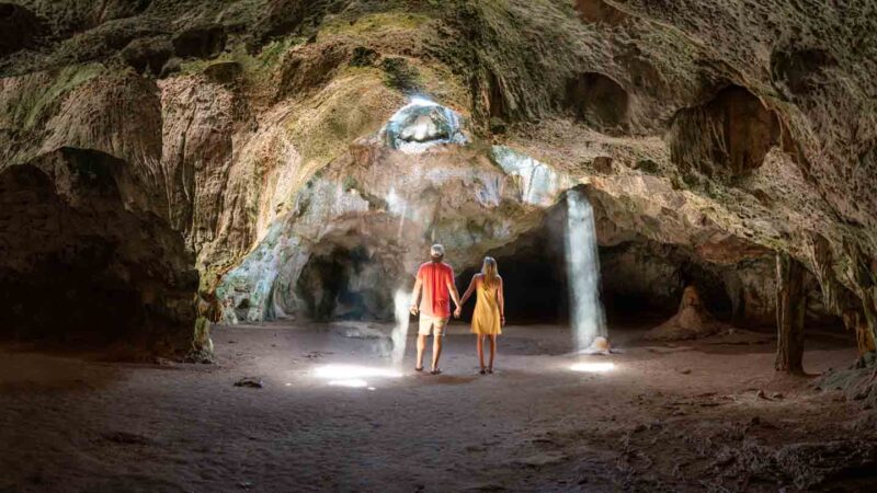 Pareja de pie dentro de la cueva Quadirikiri Aruba con rayos de luz que atraviesan el techo de la cueva Aruba