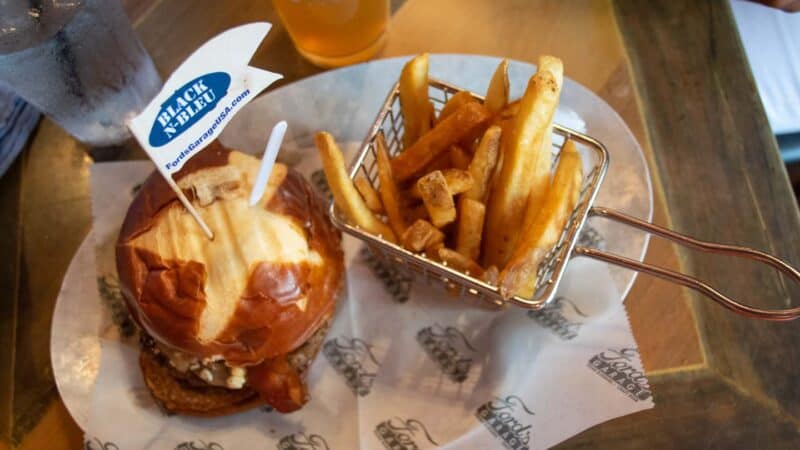 a large burger with a white and blue flag next to a wire basket of french fries served at Fords Garage Restaurant in Ft Myers