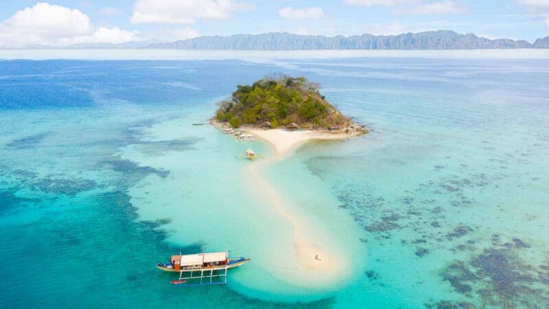 Drone photo of a traditional boat anchored in aqua colored waters next to a thin strip of sand connected to Bulog Dos Island Coron