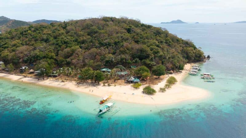 Aerial view of Banana Island with boats, people swimming, large beach, and small huts to stay