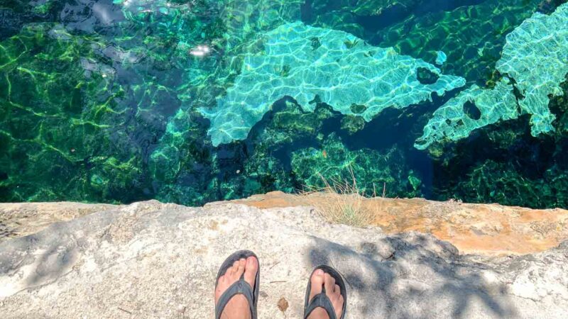 Man's feet standing on limestone cliff looking down into the blue waters of Cenote Azul