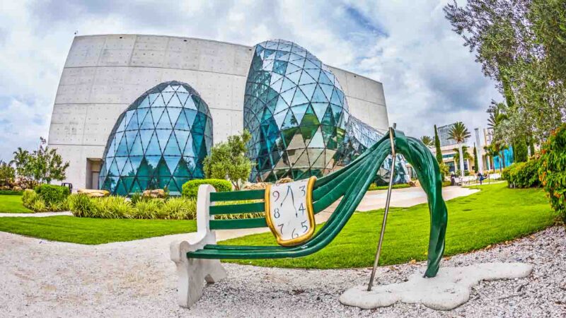a green bench warped with a melting clock in Salvador Dali style in front of the concrete and glass Dali Museum in St. Petersburg Florida
