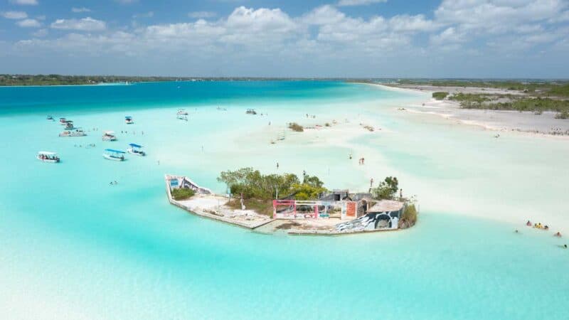 Drone photo of Canal de Los Piratas filled with colorful water (blues and greens) looking back at the city of Bacalar Mexico in the distance