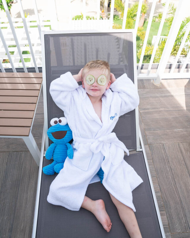 Toddler boy in a rob with cucumbers on his eyes in a lounge chair with Cookie Monster stuffed animal 
