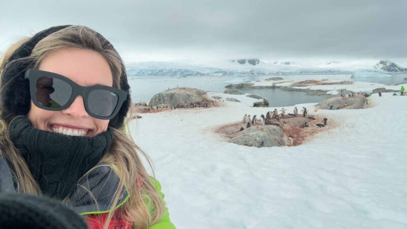 woman with pengiuns in Antarctica wearing neck gator, sunglasses, and earmuffs