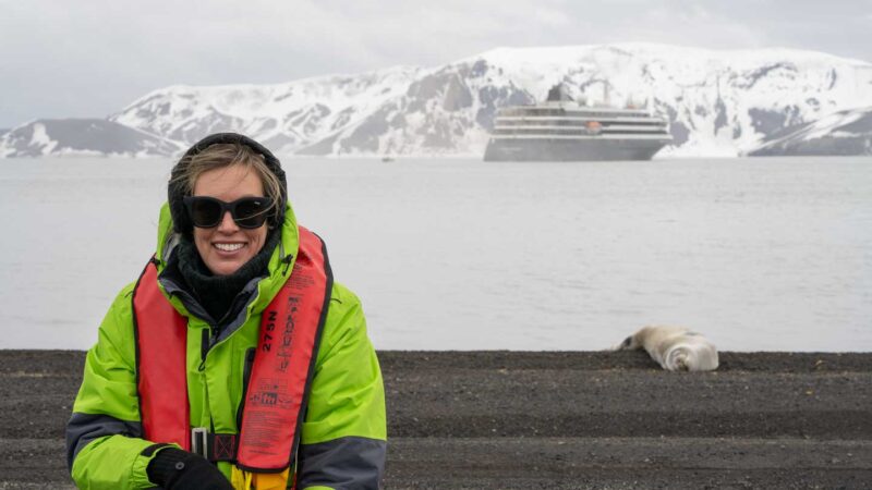 Woman in Antarctica with a seat and Atlas Ocean Voyages World Voyage cruise ship behind her. 