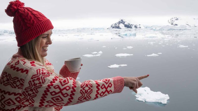 woman pointing to an iceberg in Antarctica wearing a hat & sweater, holding a cup of coffee