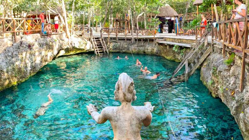 Stone statue overlooks Zacil Ha Cenote with clear blue water and people swimming - One of the best cenotes near tulum